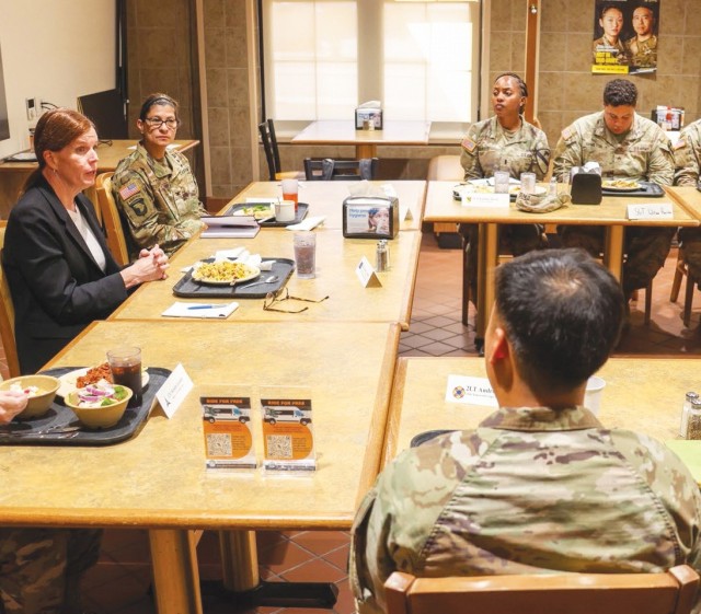 People sit at table put together to have them face each other with trays of food in front of each person. One person at the head talks. 