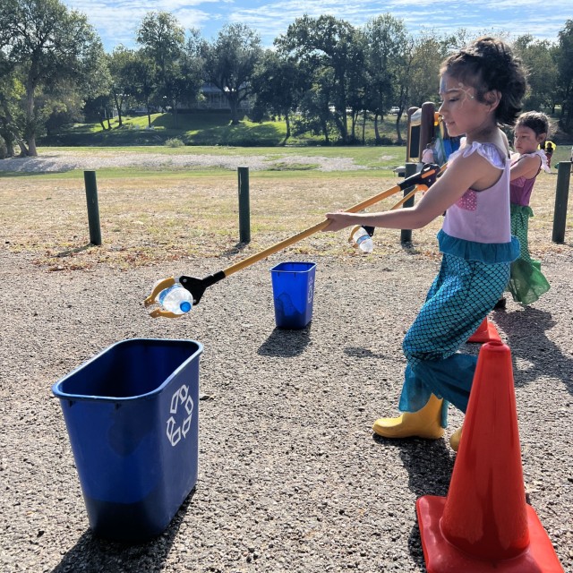 Two kids holding long grabber tools with plastic water bottles clamped in them move toward blue recycling bins. 