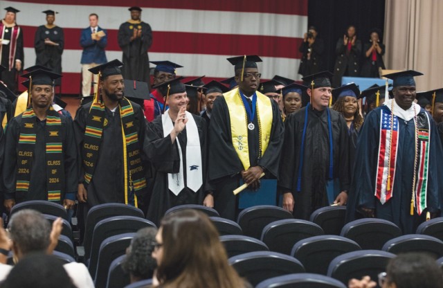 Rows of people standing and dressed in graduation caps and gowns face chairs as people look on and clap.