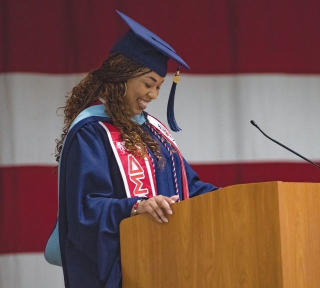 A woman smiling down and dressed in a graduation cap and gown stands behind a wooden podium. 