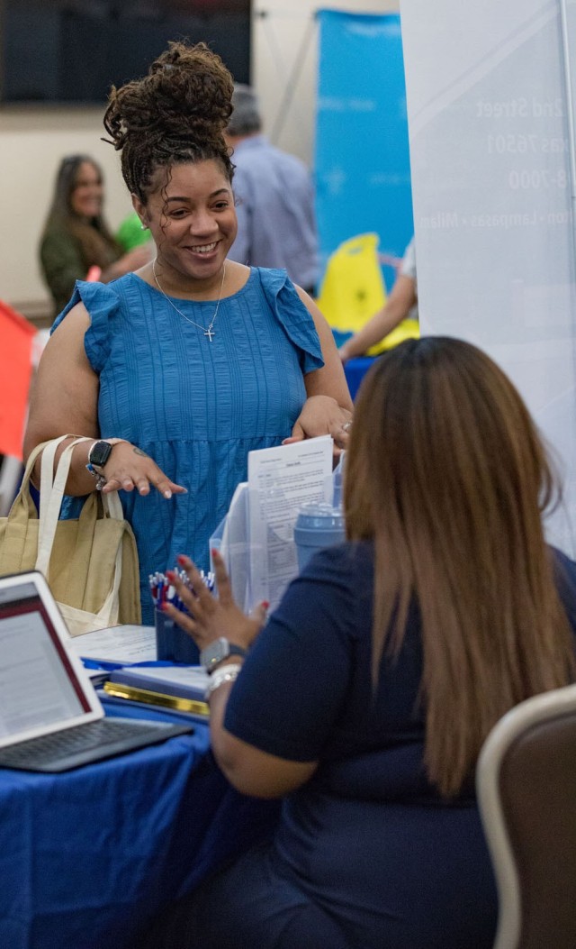 A women standing smiles as she speaks to a women sitting behind a blue clothed-covered table. 