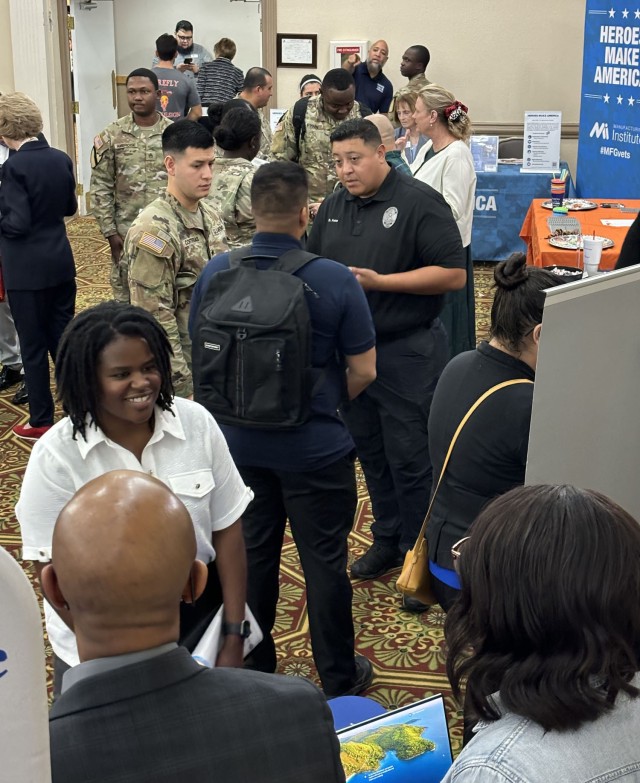 People stand looking at infographics and talking to people while sitting in front of a row of clothed-covered tables with freebies and informational papers sitting on top of them. 