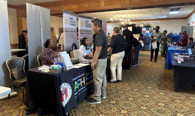 People standing talk to people sitting behind a row of clothed-covered tables with freebies and informational papers sitting on top of them. 