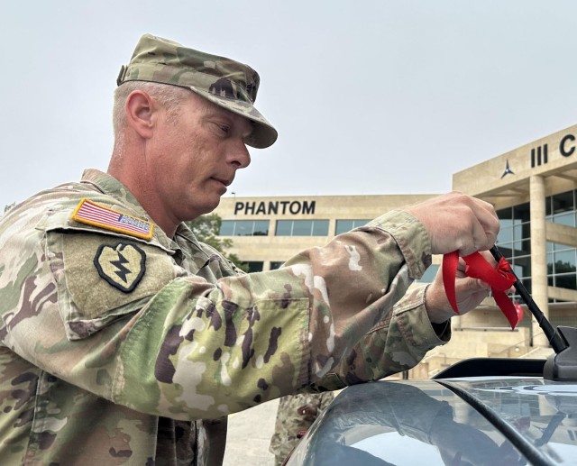 A man dressed in an army combat uniform ties a red ribbon to the antenna atop a motor vehicle.