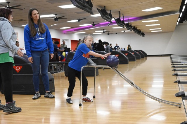 A young girl pushes a bowling ball off a bowling ramp into a lane as two adult women stand, looking on. 