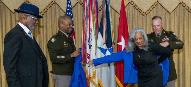 A man standing assists a woman in putting on a blazer as two men to their right look on. 