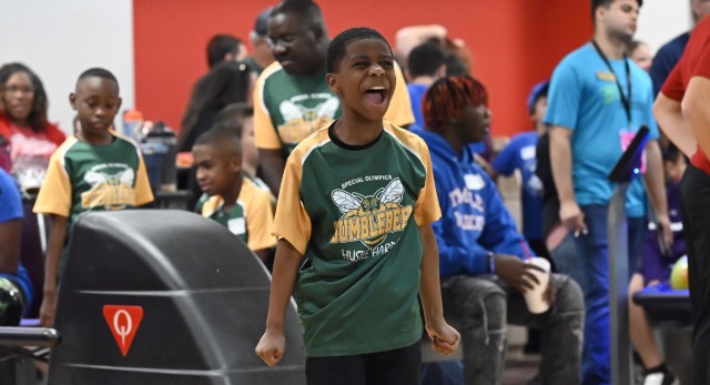 A young boy cheers, mouth open wide with his arms down by his sides and hands in fist, standing as people mill about in the background.