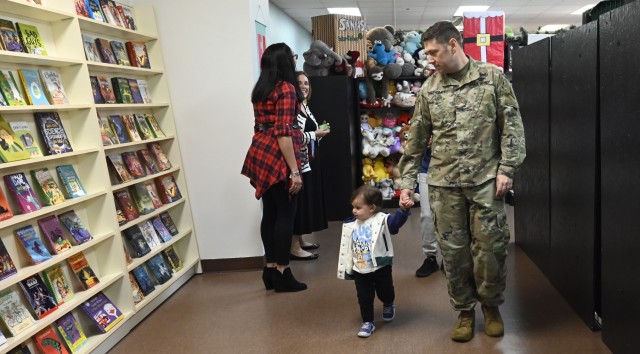A man stands hand in hand with a young boy, both looking at books displayed on white shelves.