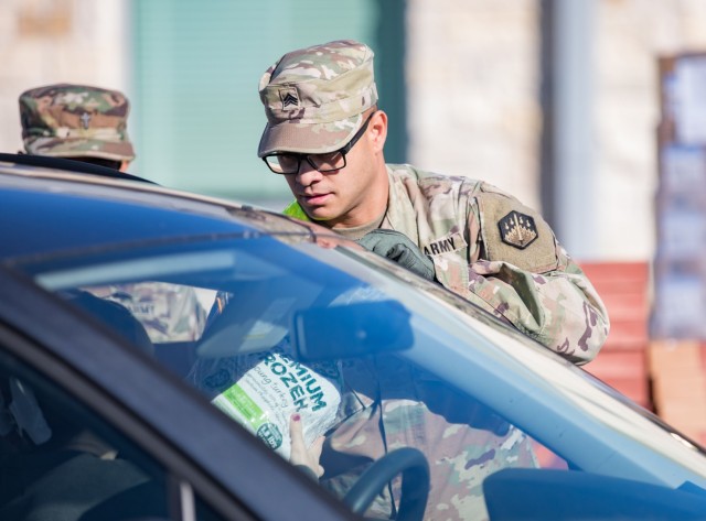 A man standing and dress in an Army combat pattern uniform hands a frozen turkey to a woman sitting in the driver&#39;s seat of a vehicle through an open window.