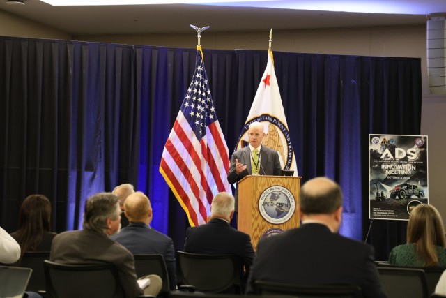 Man stands at podium in front of two flags as he give remarks to an audience of business professionals
