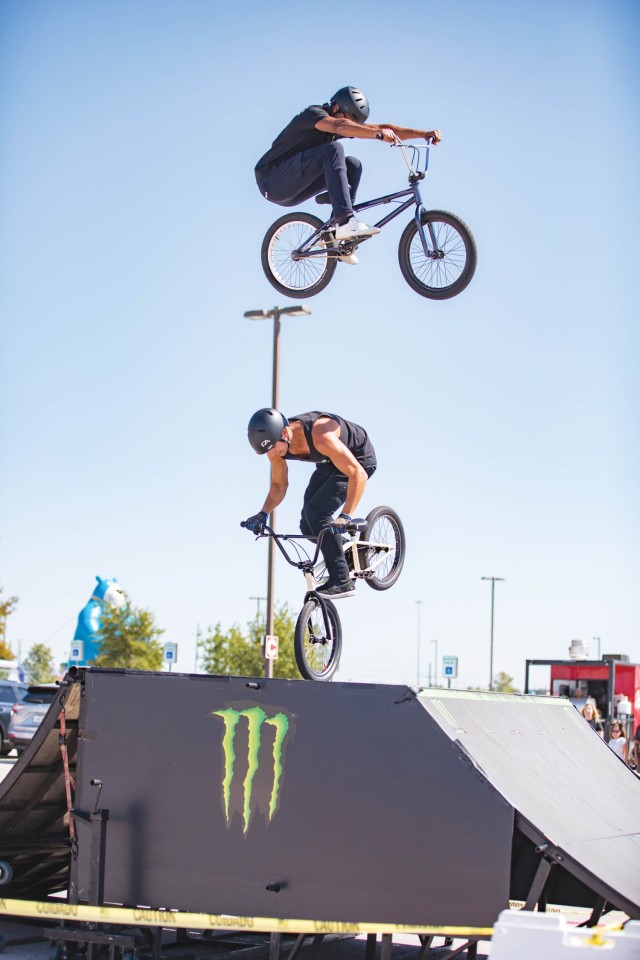 A man is in the air mounted on a bike. Straight below him, another man on a bike balances the bike on its front wheel.