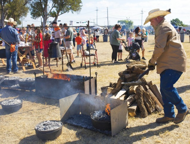 A man looks down at a coal fire in a metal bowl while standing by a pile of chopped firewood. Past him, people gather around a table.