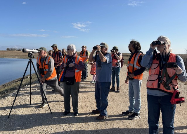 A group of people wearing bright orange vests over t-shirts look through scopes, some handheld and one on a tripod. 