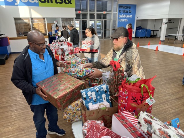 People stand and hold wrapped, rectangular boxes around a row of tables piled with wrapped box and bags with tissue paper. 