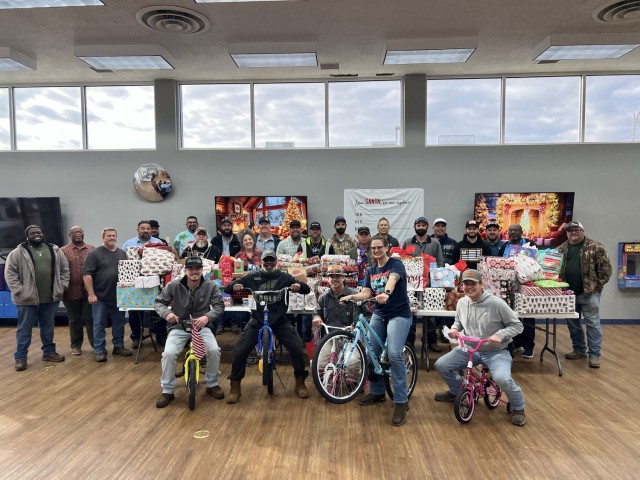 A group of people pose for a people, a majority of the people standing close together behind a connected row of tables piled with wrapped gifts and some people sitting atop bikes in front.