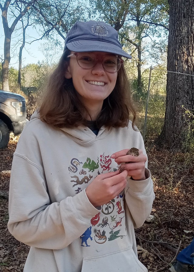 A woman standing and smiling poses for a photo with a small, brown bird held in her left hand.