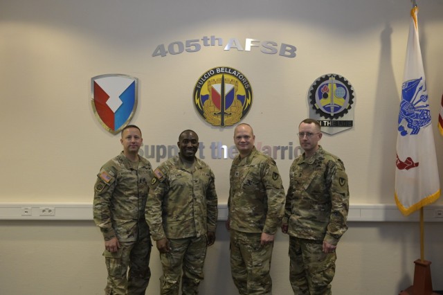 Four people pose for a photo in front of a wall with logos on it. They all wear military uniforms.