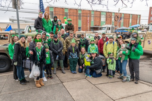 Several people pose for a photo in front of military vehicles. Most are dressed for St. Patrick&#39;s Day.
