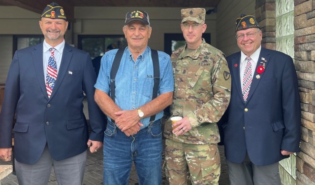 Four people pose for a photo in an outdoor area. One wears a military uniform.
