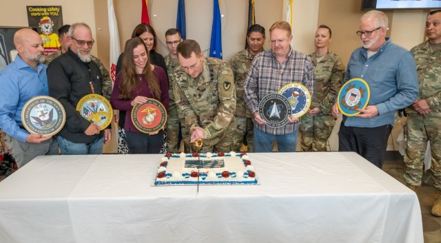Several people pose behind a table and look on and hold the seals of U.S. military branches as one person uses a sword to cut a cake.