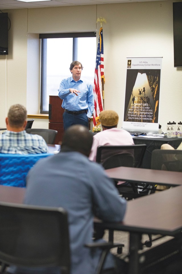 A man standing in front of a room of people sitting, talks while gesturing with right hand. 