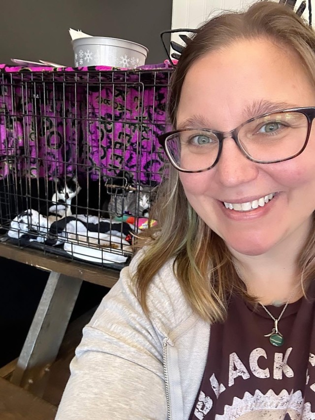 A person poses for a selfie with several cats in a wire crate in the background.