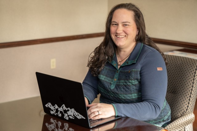 A person sits at a table in a conference room with a laptop in front of them. The person looks at the camera and smiles.