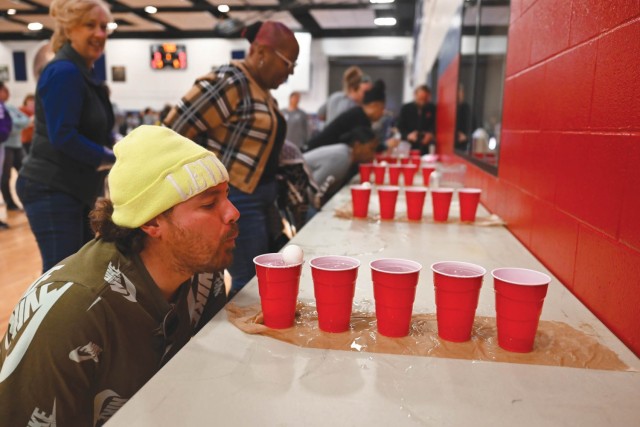 A man squats in front of a table flush against the wall lengthwise, blowing a ping pong ball floating on water in a red solo cup in a row of other water-filled red solo cups.