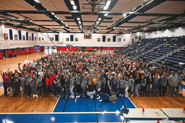 Approximately 700 people stand in a gymnasium, grouped together in a uniform rectangle to pose for a photo.