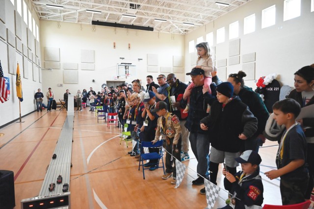 Adults and children stand behind a line with triangle checkers flags hanging along it held up by chairs, looking at four decorated, wooden cars along a metal, four-lane racetrack in an open room.
