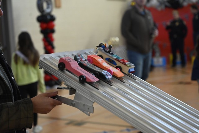 Four decorated wooden cars sit on a metal, four-lane racetrack as someone standing holds a level on the side of the metal track.