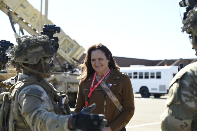 A woman standing smiles as she looks at men dressed in an Army combat pattern uniform with tactical gear and paint atop.