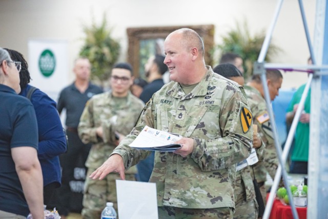 A man standing and dressed an Army combat pattern uniform gestures with his right hand while holding a stack of papers in the other, looking at a woman across from him.