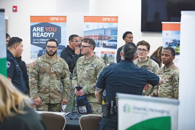 People standing and dressed in Army combat pattern unforms looks at two men in black uniforms standing behind a black-clothed table.