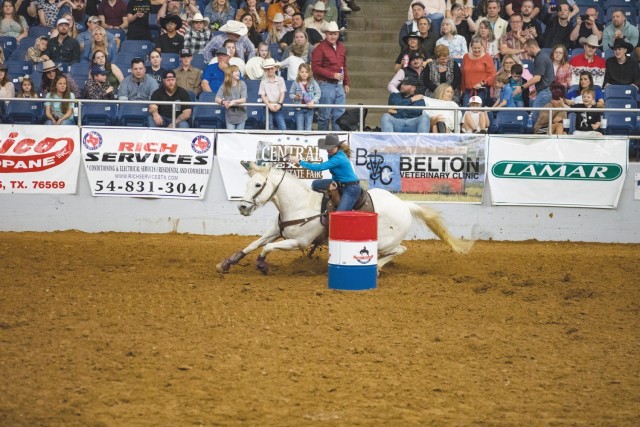 A woman sits on a saddle on a horse holding onto reins as the horse goes a around a multi-colored barrel as people sitting in stands look.
