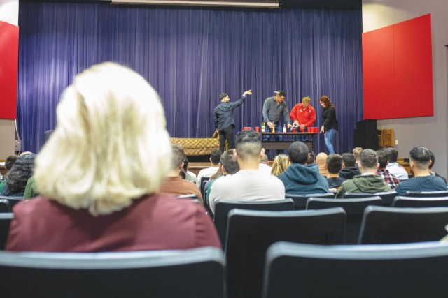 People sit in auditorium chairs watching as people on an elevated stage gather around a table with several red plastic cups. 