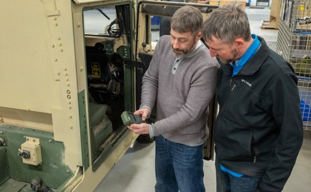 One person holds a piece of equipment as a second person looks on. They are standing near a military vehicle.
