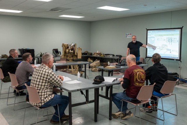 Several people sit around tables in a classroom as an instructor teaches.