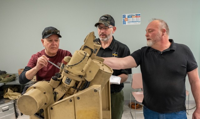 Three people look at a piece of military equipment. One instructs and the other two listen.