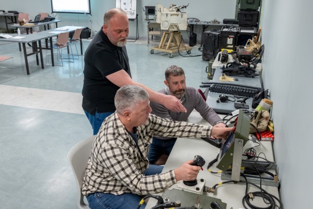 Three people are pictured. One gestures toward a piece of military equipment, one trains on the equipment, and one more looks on.