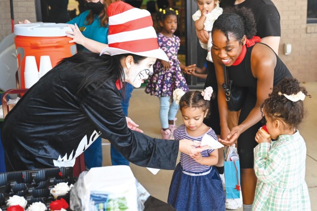 A woman with face painting, a black, long-sleeved shirt and a tall red and white striped hat hands a cupcake to a woman and two young girls.