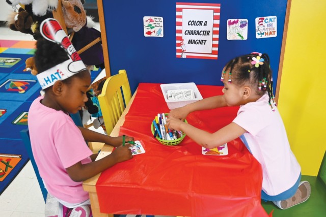 Two young girls sit across from each other at a table, one girl using a marker on a piece of paper while the other sifts through a basket of markers.