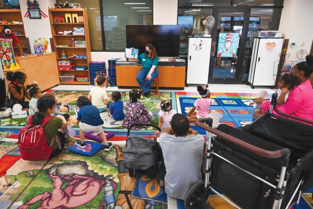 A woman sitting in a chair holds a book open in her right hand as a group of children sit on rugs and look on.