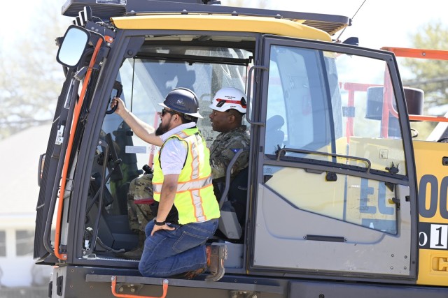 A man sits in the cab of a large machine while another crouches on a step just outside the cab, holding onto to a handle in front of him.