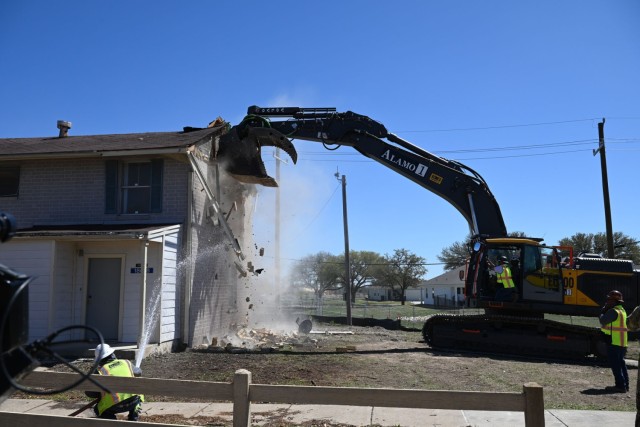 An excavator uses the arm to push against a house, destroying it with debris raining to the ground while a man aims a running water hose at the falling debris.