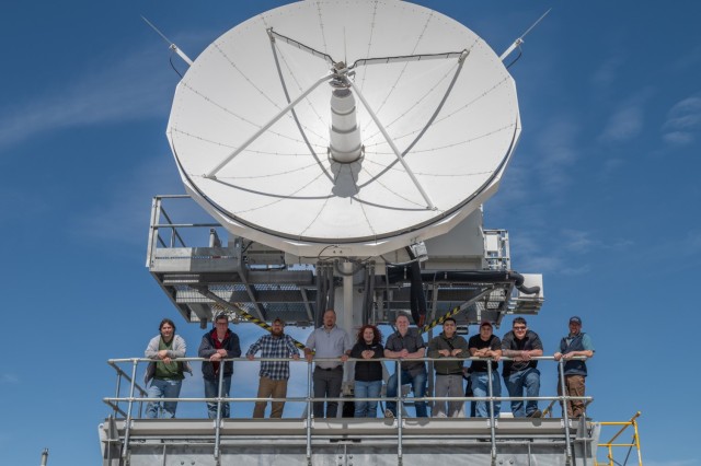 Several people pose for a photo near a piece of satellite communications equipment.