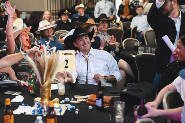 A man sitting at a rounded table holds up a paddle with a laminated piece of paper attached to it with the number two one. On his right, a woman raises her right hand in the air, her mouth wide open.