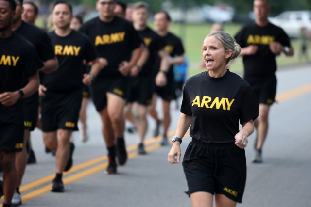 An Army Soldier with a black Army shirt and shorts calls cadence as other Soldiers run in formation.