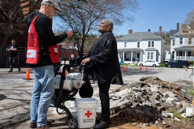 A man speaks to a woman who has a wheeled walker filled with paper towels and a Red Cross white bucket sitting next to her.