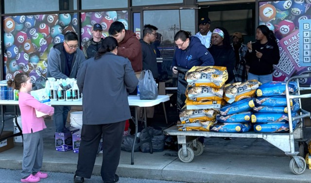 Several people stack dog food and bottles of cleaner onto a white table and trolly while a woman and her child look at the supplies.